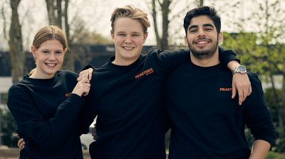 Tre elever i arbetskläder som ler och håller armarna om varandra utomhus.