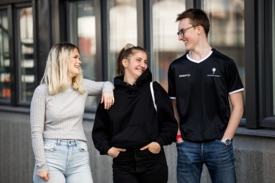 Tre elever utanför skolans lokaler som står och skrattar i solen