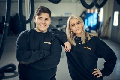 Två elever som står nära varandra i verkstaden och tittar mot kameran.