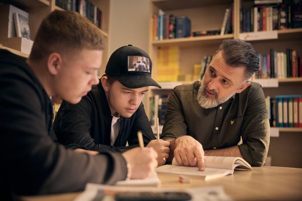 Två elever som pluggar i biblioteket tillsammans med en lärare.