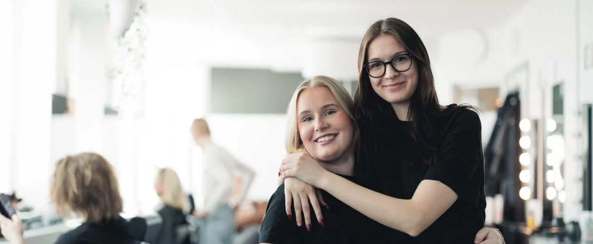 Två elever i ett klassrum håller om varandra och kollar in i kameran