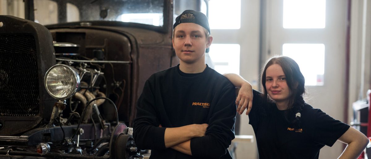 Två elever som mekar med en bil står och kollar in i kameran