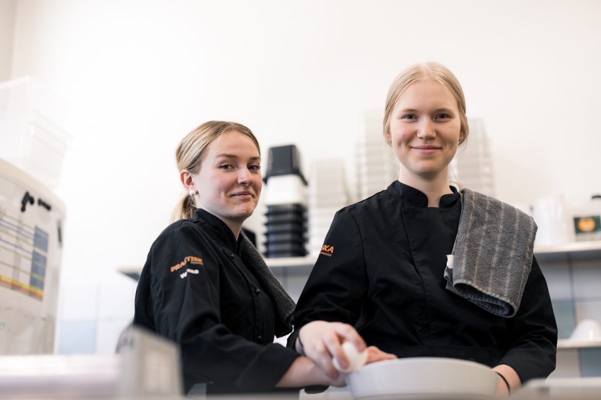 Två elever i kockkläder i ett kök tittar in i kameran
