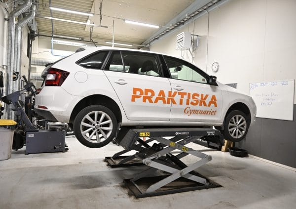 En vit bil med texten "Praktiska Gymnasiet" som är upphöjd på en ramp i en bilverkstad.