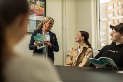 Tre elever på Hotell och turism lyssnar på en lärare som håller i kurslitteratur om besöksnäring och turism.